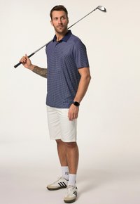 Námořnické polo tričko s potiskem, bílé šortky a tenisky s béžovými akcenty. Golfová hůl opřená na rameni, hodinky na zápěstí.