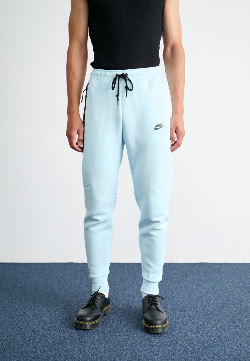 Light blue sweatpants with a drawstring waist, side pockets, and black Nike logo; paired with black lace-up shoes on a blue carpet.