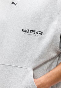Sudadera sin mangas Puma de color gris claro con bolsillo frontal, logo Puma negro y texto "PUMA CREW '48" en el pecho, mano apoyada en el bolsillo.