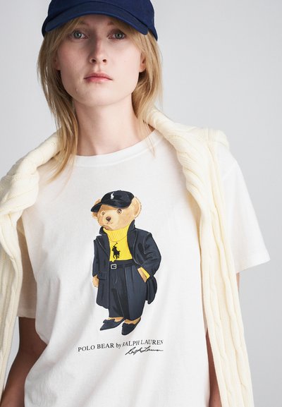 T-shirt blanc arborant un ours de dessin animé portant un manteau noir et une chemise jaune, avec une casquette noire, se tenant de manière confiante. Un cardigan en tricot léger drapé sur l'épaule.
