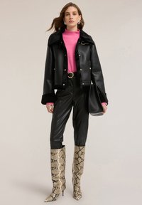 Donna che indossa una giacca nera in ecopelle con bordatura in pelliccia, maglione rosa, pantaloni neri, stivali in pelle di serpente e tiene una borsa nera.