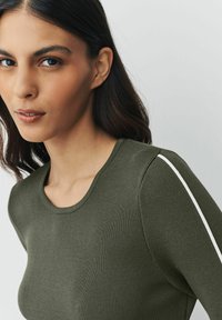 Olijfgroen geribd langslavenshirt met een ronde halslijn en witte strepen langs de schouders. Glad materiaal, aansluitend ontwerp.