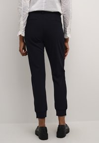 Marinblå nylånga byxor i mjukt, stretchigt tyg, med elastisk midja, sidofickor och dragkedjor vid vristerna; matchas med en vit skjorta med knappar.