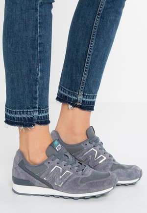 Personne portant des baskets New Balance grises et un jean bleu foncé court avec des bords effilochés, debout sur une surface blanche.