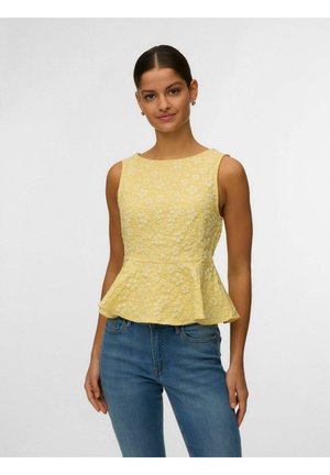 Kobieta w żółtym, kwiecistym, teksturowanym topie peplum bez rękawów, w połączeniu z niebieskimi dżinsami, stojąca na tle jednolitym, jasnym.