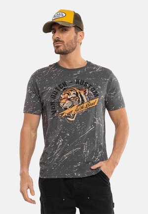 Homme portant un t-shirt gris usé avec un tigre rugissant et le texte « Von Dutch Hustman, Fuel the Beast », ainsi qu'une casquette jaune et marron.
