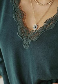 Gros plan sur un haut en maille vert foncé avec un décolleté en V en dentelle festonnée et des colliers dorés superposés, dont un avec un pendentif en forme d’étoile.