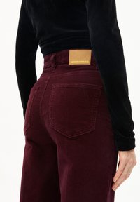 Bordeaux kleurige corduroy broek met een leren label op de tailleband, voorzien van twee achterzakken en een textuurpatroon. Zwarte top gedeeltelijk zichtbaar.