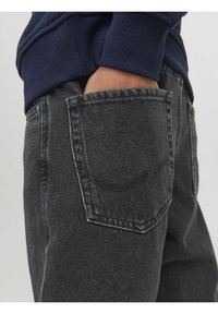 Jeans de denim gris oscuro con un bolsillo trasero que presenta una costura curva sutil y un detalle de botón azul cerca de la cintura. La tela tiene una textura clásica.