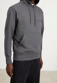Šedá mikina s kapucí a předním kapsou, šňůrkovou kapucí a logem Calvin Klein na hrudi. Vyrobena z měkké, texturované látky.