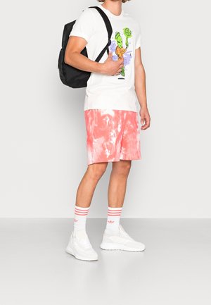 Bílé tričko s barevným motivem mimozemšťana. Lososově růžové šortky s batikováním, bílé tenisky s texturovanými detaily a pruhované bílé ponožky. Černý batoh.