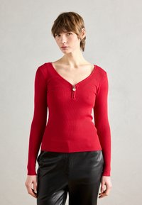 Haut à manches longues rouge côtelé avec un décolleté en V profond et un zip à l'avant. Porté avec un pantalon en cuir noir brillant.