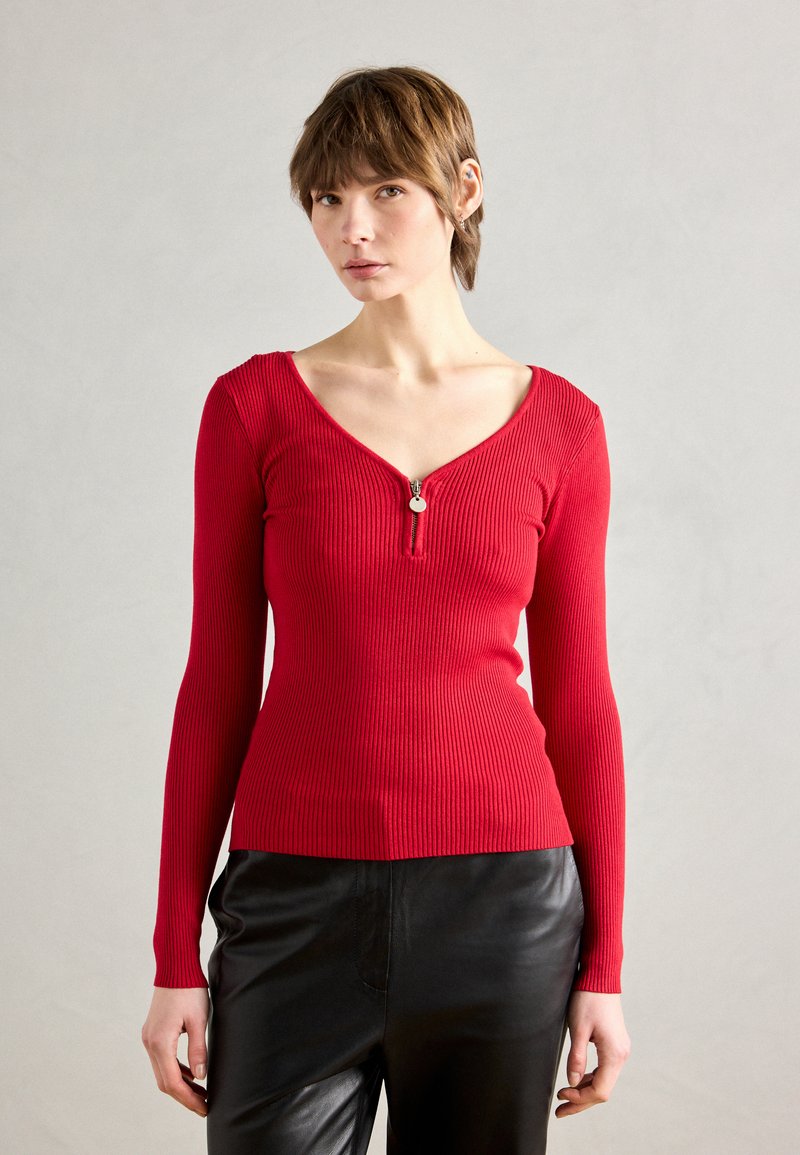 Haut à manches longues rouge côtelé avec un décolleté en V profond et un zip à l'avant. Porté avec un pantalon en cuir noir brillant.