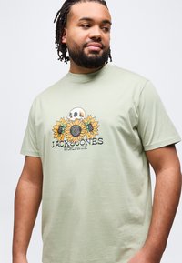 Άντρας με γενειάδα και dreadlocks που φοράει ανοιχτό πράσινο T-shirt με γραφικό σχέδιο κρανίου και ηλιοτροπίων και το κείμενο «Jack & Jones Worldwide».