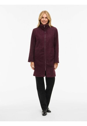 Bordeaux fleecejas met hoge kraag en ritssluiting aan de voorkant, met lange mouwen en zijzakken, gedragen over zwarte broek en schoenen.