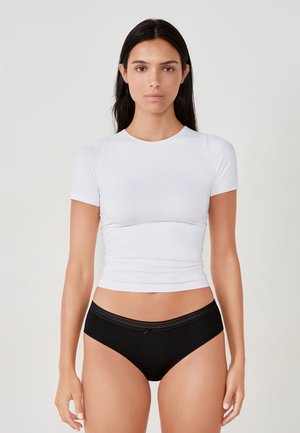 Mujer con cabello largo y oscuro, vestida con una camiseta blanca ajustada de manga corta y ropa interior negra, de pie frente a un fondo claro y liso.