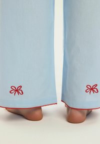 Jasnoniebieskie spodnie z haftem czerwonej kokardy i czerwoną koronką na brzegu, noszone przez osobę stojącą boso na jasnej powierzchni.