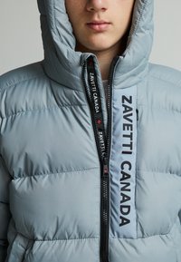 Veste matelassée bleu clair avec une capuche. Présente des coutures horizontales, une fermeture éclair sur le devant avec un tirette noire, et "ZAVETTI CANADA" imprimé en évidence.