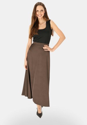 Uśmiechnięta kobieta z długimi włosami, ubrana w czarny bezrękawnik, brązową asymetryczną spódnicę z wysokim stanem oraz czarne spiczaste buty, stojąca z ręką na biodrze.