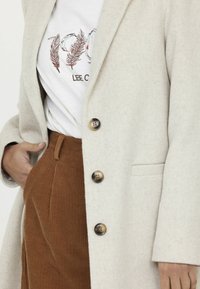 Hellbeige Wollmantel mit vier dunklen Knöpfen, kombiniert mit braunen Cordhosen. Das T-Shirt zeigt rote und braune Federstickerei.