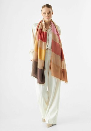 Longue écharpe rayée aux teintes chaudes de rouge, rose, jaune et marron. Fabriquée en tissu doux avec des bords frangés, superposée sur une tenue crème.