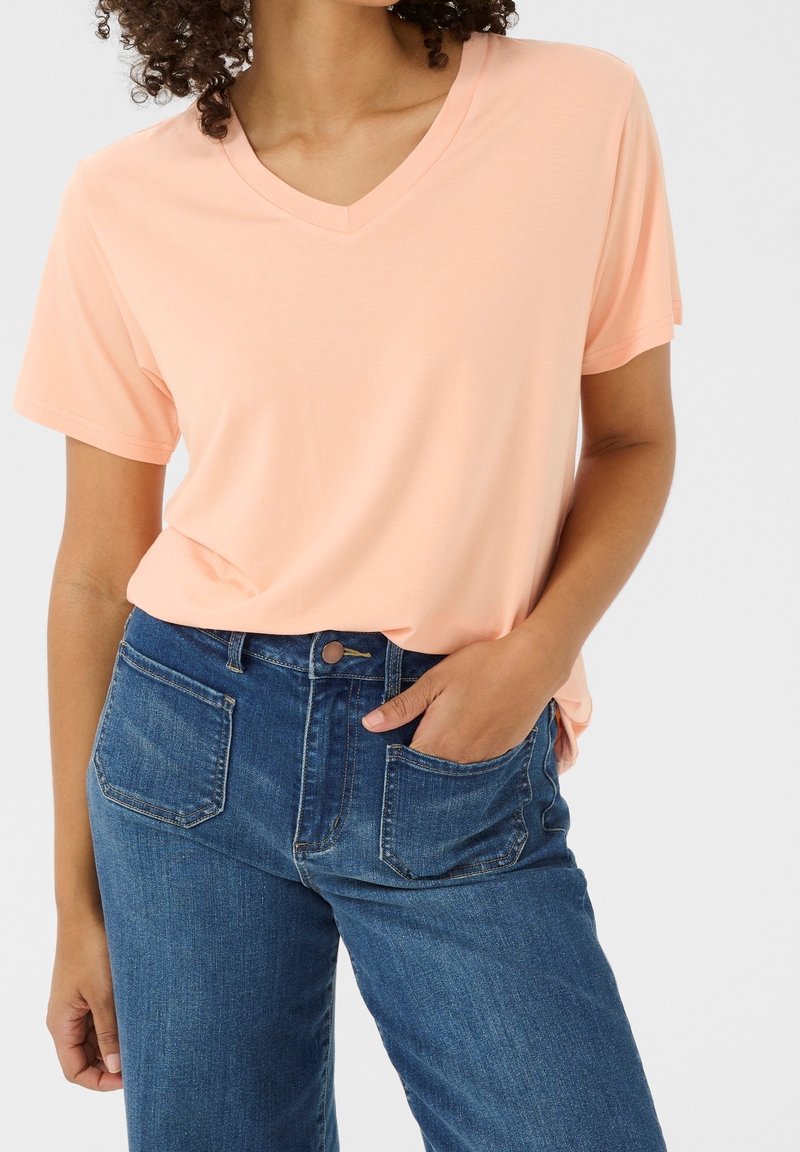 Femme portant un t-shirt à manches courtes couleur pêche clair avec un col en V, rentré dans un jean en denim bleu avec des poches plaquées à l'avant, une main dans la poche.