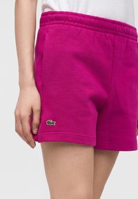 Femme portant un short rose vif avec une ceinture élastique et un petit logo crocodile vert près de l'ourlet gauche.