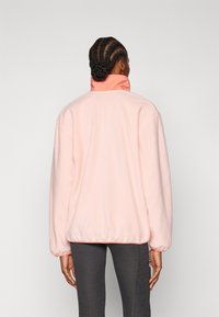 Vaaleanpunaista fleece-takkia, jossa on korkea kaulus, elastiset hihansuut ja helma. Pehmeä rakenne, jossa on ompeluyksityiskohtia reunojen ympärillä.