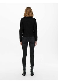 Veste en denim noir à coupe slim, manches longues et poignets à boutons. Portée sur un jean skinny noir et des bottines, vue de dos.