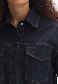 Chemise en denim foncé avec deux poches poitrine, coutures orange visibles, col et fermeture à boutons. Le tissu semble solide et texturé.