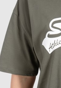 Camiseta de manga corta en verde oliva de algodón suave con cuello redondo y logo blanco impreso en la parte delantera. Detalles de costura visibles.