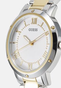 Silver and gold-tone watch with a white dial, Roman numeral markers, and three-hand movement. Features a metal and silicone bracelet.