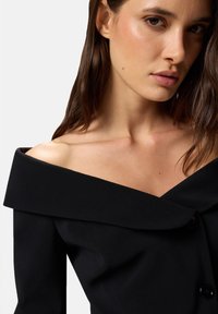 Abito nero scollo a barca con ampio colletto, corpetto aderente e dettaglio con bottoni sul petto. Tessuto liscio con una leggera lucentezza.