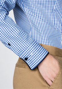 Manga de camisa a cuadros azul y blanca, con botones negros y ribete azul marino, combinada con pantalones caqui.