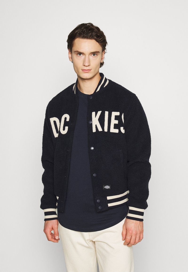 Giacca varsity nera in fleece con lettering crema. Dotata di polsini e colletto a coste rigate e chiusura con bottoni.