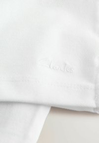 Vit tyg med en broderad "Clarks"-logotyp i en subtil, texturerad finish syns i förgrunden och visar på dess kvalitet.