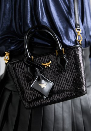 Fekete kézitáska flitteres textúrával, sima fülekkel, arany fémszegélyekkel és díszes poggyász címkével. Kompakt kialakítás és strukturált forma.