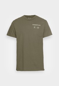 Camiseta de algodón color verde oliva con mangas cortas, cuello redondo y estampado frontal que presenta "PRIMITIVE" y detalles del logotipo en blanco.
