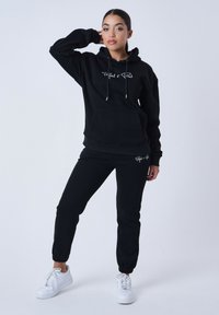 Zwarte hoodie met een voorzak en aantrekkoorden, gecombineerd met bijpassende zwarte joggers. Witte sneakers maken de outfit compleet.