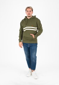 Felpa verde oliva con strisce orizzontali bianche, tasca a marsupio, cappuccio con cordino e logo ricamato, abbinata a jeans blu e sneakers bianche.