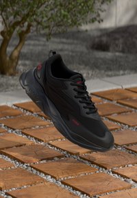 Svart sneaker med glatt materiale, avrundet design, snøringssystem og røde logoakenter. Ligger litt hevet over en teksturert overflate.