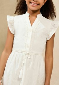 Weißes Baumwollkleid mit kurzen, gerüschten Ärmeln, durchgehender Knopfleiste und gewelltem Kragen. Mit Spitzenmuster und tailliertem Bund mit Fransen.