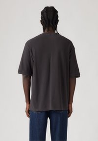 Tmavě šedé oversized tričko s kulatým výstřihem, krátkými rukávy a hladkou texturou. Kombinováno s modrými džíny.