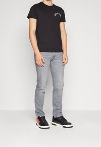 Camiseta negra de algodón con el logo "HILFIGER" en blanco en el pecho, combinada con jeans gris claro y zapatillas negras con detalles en naranja.