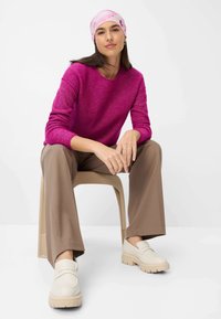 Fuchsiastrickpullover, braune weit geschnittene Hose und cremefarbene Slipschuhe mit dicken Sohlen. Das Model trägt ein gemustertes, hellrosa Kopftuch.