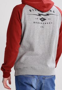 Sudadera gris con mangas raglán rojas, que presenta un logo impreso en la parte posterior, cinturilla elástica y textura de jersey suave. Diseño casual para mayor comodidad.