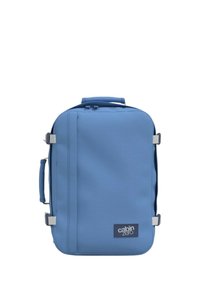 Zainetto blu con un tessuto liscio e resistente, forma rettangolare, manico superiore per il trasporto, cinturino laterale e una patch con logo nella parte inferiore.