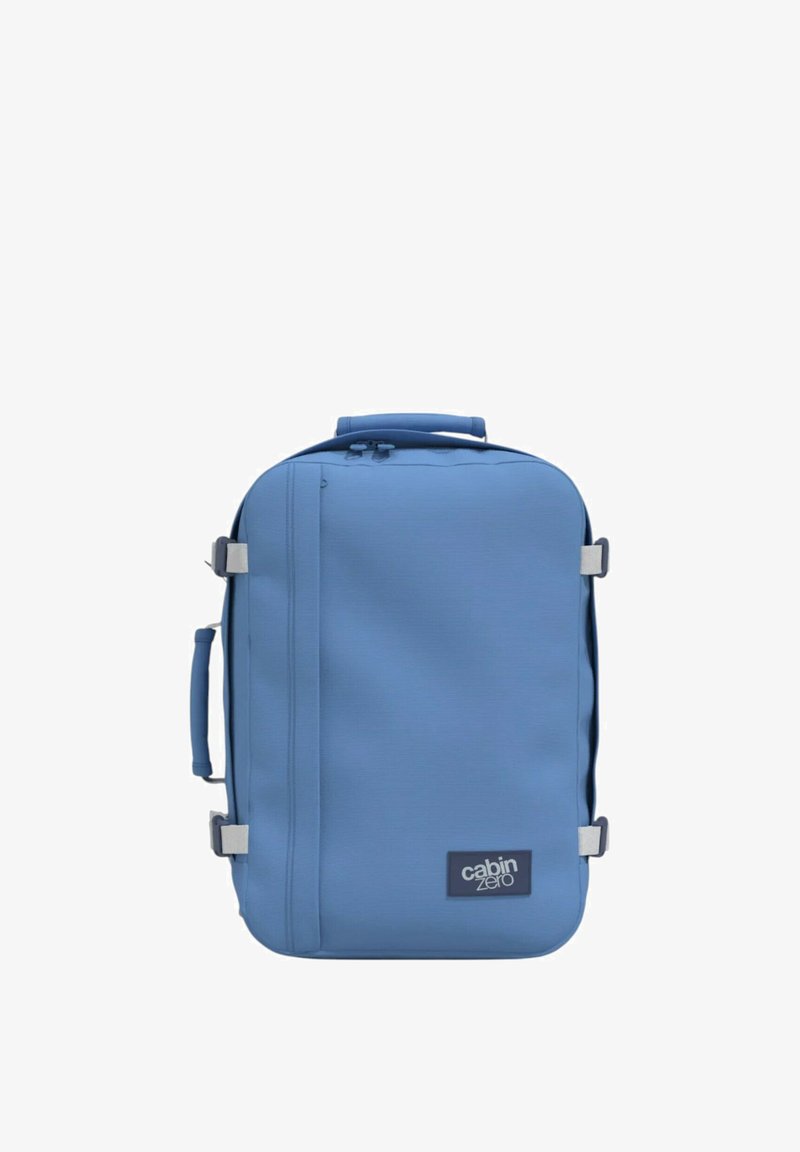 Zainetto blu con un tessuto liscio e resistente, forma rettangolare, manico superiore per il trasporto, cinturino laterale e una patch con logo nella parte inferiore.