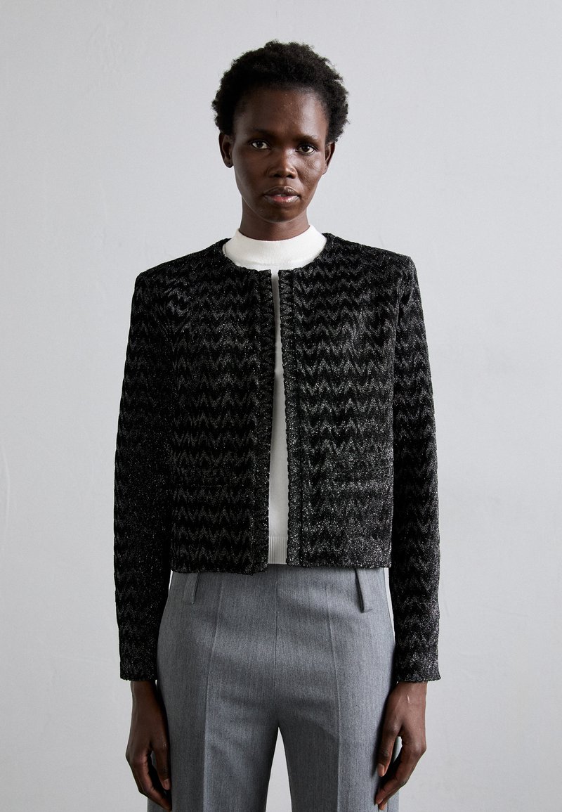 Black and silver textured jacket with a zigzag pattern, round neckline, and front closure, worn over a white sweater and gray trousers.