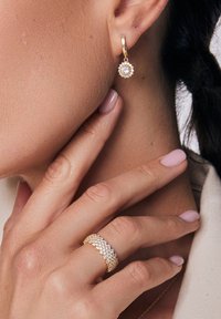 Złote kolczyki z okrągłym wisiorkiem, w którym znajduje się centralny kamień szlachetny oraz małe kamienie akcentujące. Teksturowany złoty pierścień z detalami z kamieni szlachetnych.